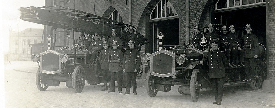 1895 bis 1920 mehrere Feuerwehrleute neben und in einer Drehleiter und ein zweiten Fahrzeug mit Feuerwehrleuten