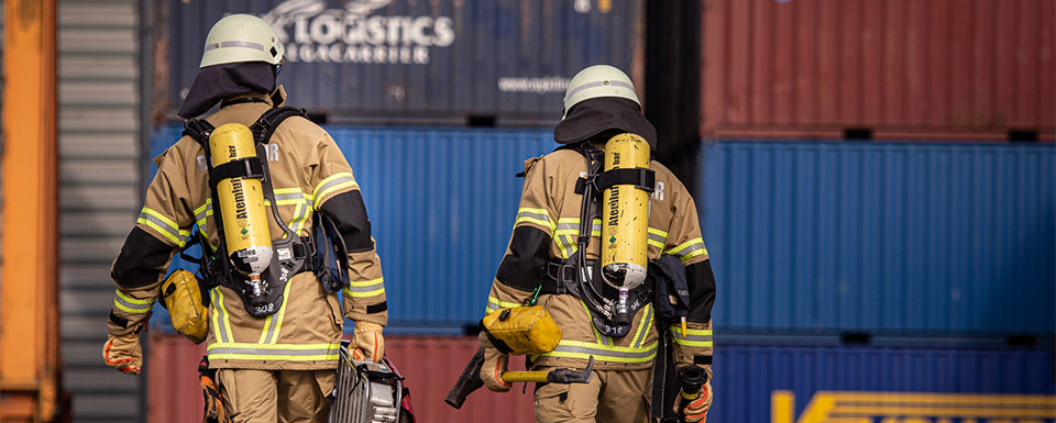 Zwei Beamte:innen in Schutzkleidung von hinten fotografiert vor gestapelten Containern