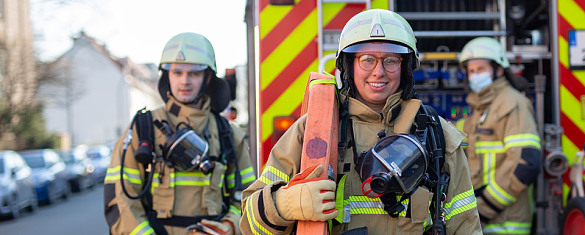 Eine Feuerwehrfrau in Schutzkleidung mittig von zwei Feuerwehrleuten hintereinem Löschfahrzeug