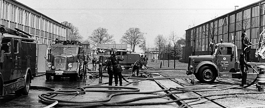 Feuerwehrübung auf dem Hof einer Feuerwache mit mehreren Einsatzfahrzeugen um 1950. Viele ausgerollte Schläuche auf dem Boden.